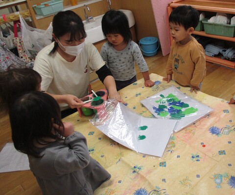 保育者が画用紙に、絵の具を載せフィンガーペイントの準備をしている様子を見ている子ども